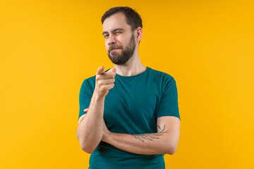 Portrait of attractive young bearded man winking and pointing with his index finger directly to the camera. Isolated over bright colored yellow background