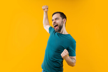 Young euphoric bearded man celebrating money win at bookmaker's after betting at favourite team, screaming yes and making winner's gesture clenching his fist. Isolated over yellow background - 476658106