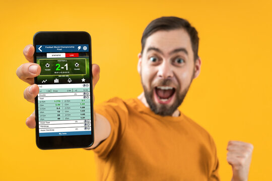 Happy excited soccer fan reaching his smartphone with bookmaker's website application interface template directly to the camera over yellow background. Focus is on the hand with phone