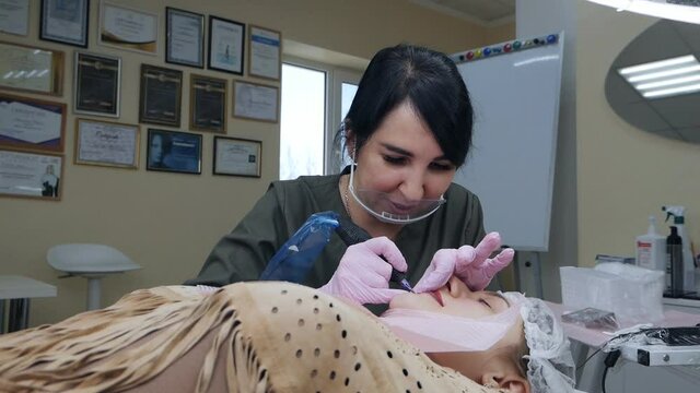 Cosmetologist applies permanent pigment tattoo to female lips with tattoo needle in salon