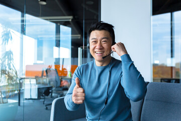 Happy asian smiling and rejoicing, businessman worker making video call, conference remotely, looking at camera and pointing thumbs up, using headset, webcam view