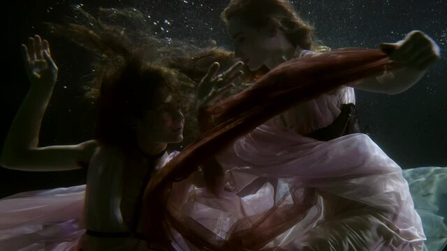 two fairy women are floating together underwater, lesbians lovers are swimming inside pool
