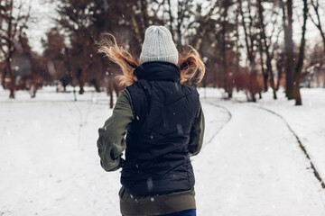Fototapeta na wymiar Young woman athlete running in snowy winter park under falling snow. Back view. Active healthy lifestyle
