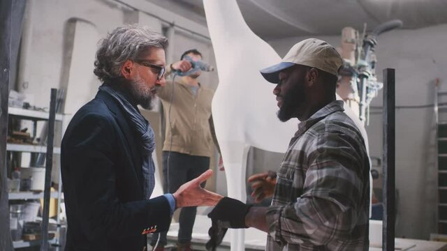 African American craftsman and mature customer shaking hands while striking deal near colleagues making giraffe statue in professional creative workshop