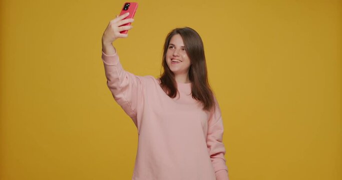 Selfie portrait. Smiling happy young girl making selfie, posing, taking pictures using smart phone on yellow background