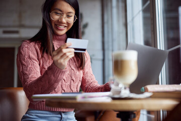 A happy Asian girl sitting in the cafeteria and using her debit card for online shopping on the laptop. E-commerce and e-banking concept.