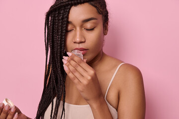 Young African woman holds a jar of a cosmetic product and smells the fragrance, posing with her eyes closed on a pink background with space for advertising text.