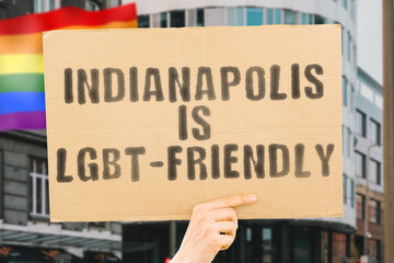 The phrase " Indianapolis is LGBT-Friendly " on a banner in men's hand with blurred LGBT flag on the background. Human relationships. different. Diverse. liberty. Sexuality. Social issues. Society