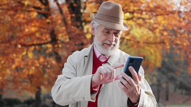 Active senior people with digital technology concept. Smiling elderly gray-haired man using smartphone for texting message,reading news,watching photo or video in social media walking in autumn park.
