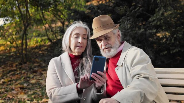Happy old mature retired couple using smartphone for watching funny photos and video, mobile applications, social media, shopping online. Middle aged spouses enjoying time together in autumn park.