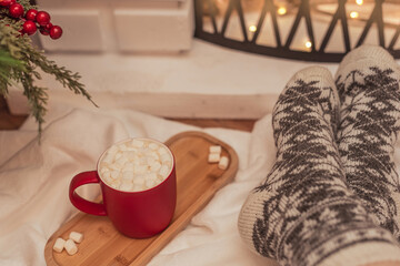 Cozy vibes scene with a cup of hot drink with white marshmallows and female feet in warm woollen socks in front of fireplace. Selective focus. Cozy atmosphere during winter holidays concept