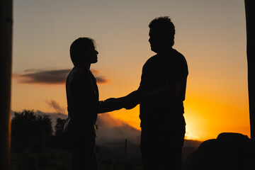 Fototapeta na wymiar Silhouette of couple at sunset with strong orange hue and mountains on the horizon and few clouds in the sky.