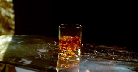A glass of whiskey breaks a glass surface in super slow motion, symbolic of alcoholism and the negative effects of drinking.