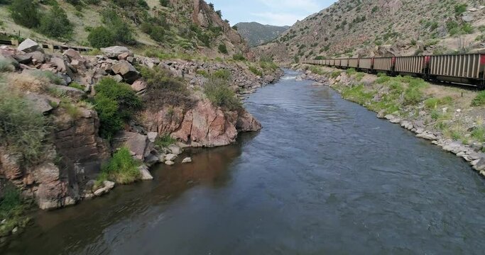 Arkansas river flowing by industrial cargo train in the Colorado Mountains during summer, aerial drone shot