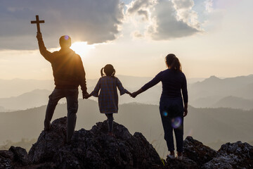 Silhouette family praying and holding Christian cross for worshipping God on mountain at sunrise...