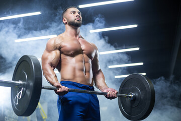 Bottom up view of a muscular athlete lifting heavy barbell showing his muscles in a gym standing in smoke