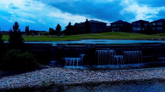 golfers play at a golf course hole surrounded by a manmade pond with waterfalls next to detached massive homes Jagare Ridge Style as clouds form on a summer afternoon in the South Edmonton Canada