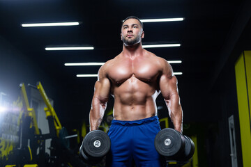 Fototapeta na wymiar Front view of a muscular bodybuilder posing with dumbbells in blue shorts in a gym