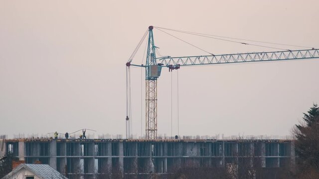 Dark silhouette of tower crane and small silhouettes of workers at high residential apartment building construction site. Real estate development