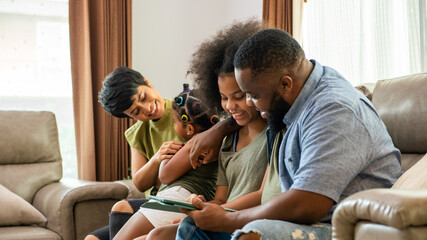 Smiling African parents and two little daughter sitting on sofa in living room using digital tablet play games or watch movie together. Happy family enjoy weekend activity with technology at homev