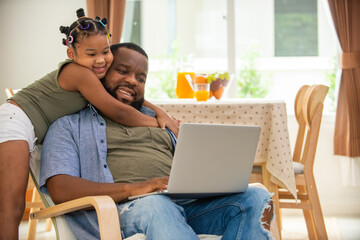 Little African daughter playing with father while he working on laptop computer on sofa in living...