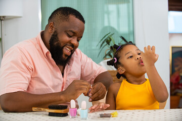 Happy family African father applying nail polish to little daughter in living room. Dad and child...