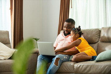 Little African daughter playing with father while he working on laptop computer on sofa in living room. Dad and child girl kid enjoy and having fun leisure activity spending time together at home