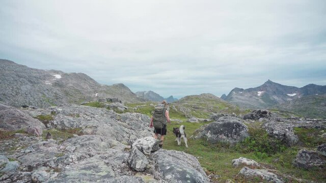 Man Backpacker With His Dog Walking On Grassy Trail With Big Rocks In Anderdalen National Park, Senja, Norway. - Wide Shot