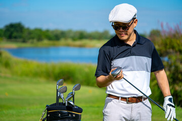 Portrait of Smiling Asian man holding golf club standing on golf course fairway in sunny day. Healthy male golfer enjoy outdoor lifestyle activity sport golfing at country club on summer vacation