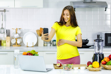 Caucasian young pregnant female mother in casual outfit with big belly tummy stand smiling holding water glass checking email from touchscreen smartphone in kitchen full of cooking equipment at home.