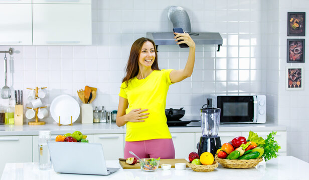 Caucasian young pregnant female mother in casual outfit with big belly tummy stand smiling holding water glass checking email from touchscreen smartphone in kitchen full of cooking equipment at home.