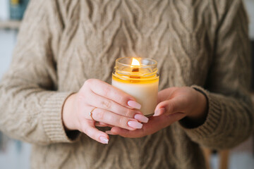 Close-up cropped shot of unrecognizable young woman holding burning scented handmade candle in glass jar near chest. Female wearing sweater holding aroma relaxing candle in hand. - Powered by Adobe