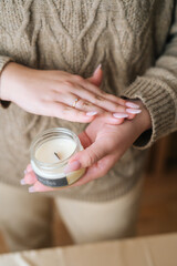 Vertical close-up cropped shot of unrecognizable young woman holding scented handmade candle in glass jar near chest. Female wearing sweater holding aroma relaxing candle in hand.
