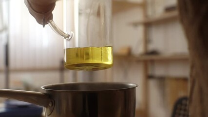 Close-up of unrecognizable female artisan pouring melted wax into glass jar over boiling pan of steam for creating candle mixture. Process of making handmade natural candle . Shooting in slow motion.