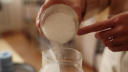 Close-up hands of unrecognizable female artisan pouring dry ingredient into glass measuring jar for creating candle building mixture. Process of making handmade natural candle. Shooting in slow motion