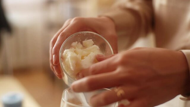 Close-up hands of unrecognizable female craftperson adding dry ingredient in glass jar into pot of boiling water for creating candle building mixture. Process of making handmade natural candle .