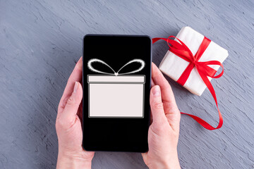 Female hands holding a phone with a gift icon and a purchased gift on a gray background. Online...