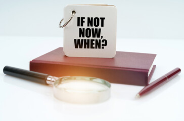 On a white surface lies a red notebook, a pen, a magnifying glass and a notebook with the inscription - If Not Now, When