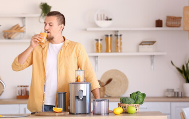 Young man with glass of fresh fruit juice near modern juicer in kitchen