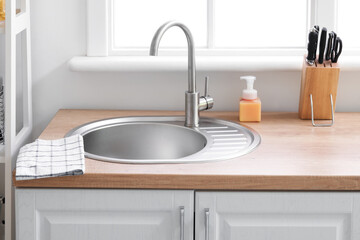 Wooden counter with silver sink, detergent and set of knives near window in kitchen