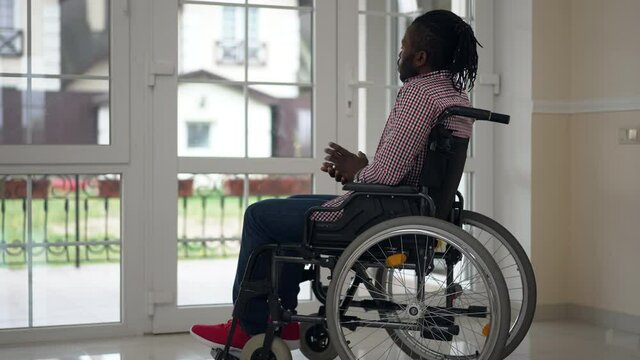 Wide shot thoughtful disabled African American man in wheelchair sitting indoors looking out the window. Portrait of young handsome paralyzed person at home in living room thinking