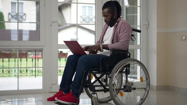 Wide shot African American disabled male freelancer typing on laptop keyboard in slow motion messaging online. Confident concentrated man working remotely from home indoors. Freelance concept