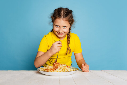 Cute little girl, emotive kid eating delicious Italian pasta isolated on blue studio background. World pasta day