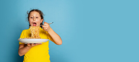 Cute little girl, emotive kid eating delicious Italian pasta isolated on blue studio background....