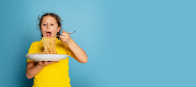 Cute Little Girl, Emotive Kid Eating Delicious Italian Pasta Isolated On Blue Studio Background. World Pasta Day