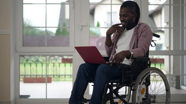 Wide shot portrait smiling disabled African American man talking using video chat on laptop sitting in wheelchair. Happy confident handsome guy messaging online at home in living room. Convenience