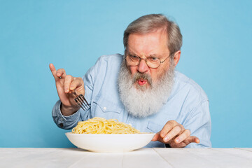 Fastidious senior hairy gray-bearded man tasting large portion of noodles, pasta isolated on blue...
