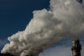 White and gray smoke and steam from a high concrete chimney against the bright blue sky