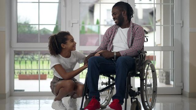 Joyful teenage girl sitting on hunkers at wheelchair with disabled African American man talking in slow motion. Portrait of relaxed happy daughter enjoying weekend leisure with father indoors