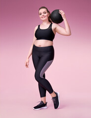 Sporty woman with medicine ball on her shoulder. Photo of model with curvy figure in fashionable sportswear on pink background. Sports motivation and healthy lifestyle